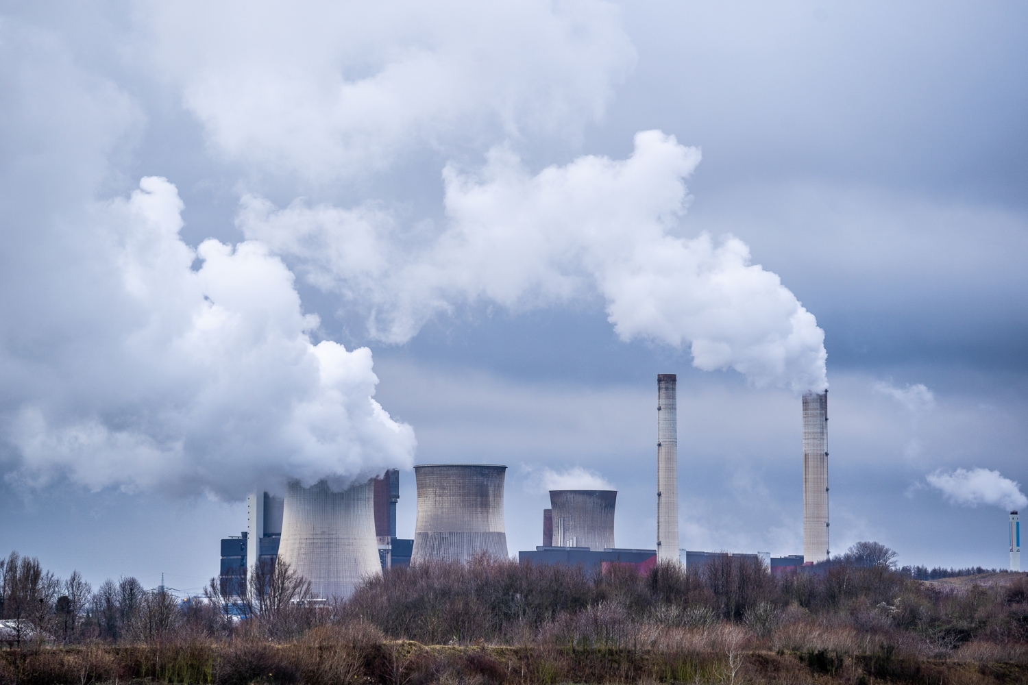 Vue d'une centrale nucléaire avec de grandes cheminées et des tours de refroidissement émettant de la fumée blanche sous un ciel nuageux